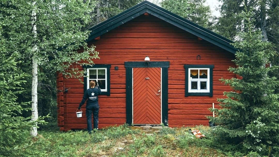 Person målar en röd träfasad på en stuga i skogen